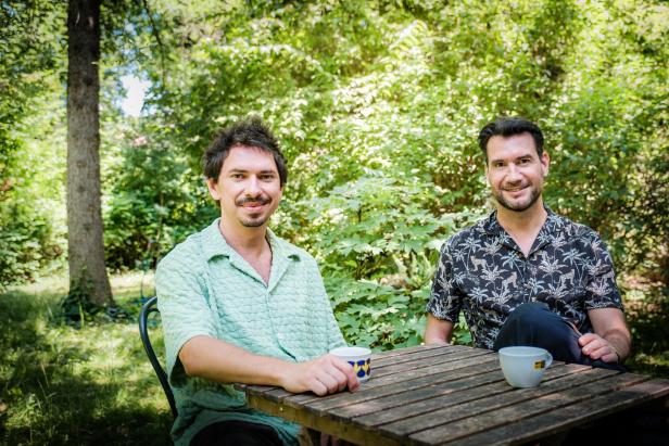 Zwei Männer sitzen an einem Holztisch im Garten und trinken Kaffee.