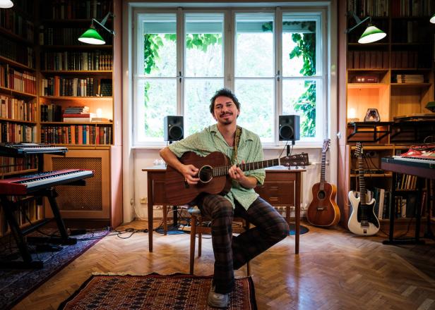 Ein Mann mit Gitarre sitzt in einem Raum voller Bücher und Musikinstrumente.
