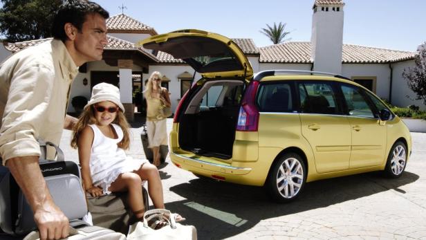 Eine Familie packt ihr Gepäck in einen gelben Citroën Picasso vor einem Ferienhaus.