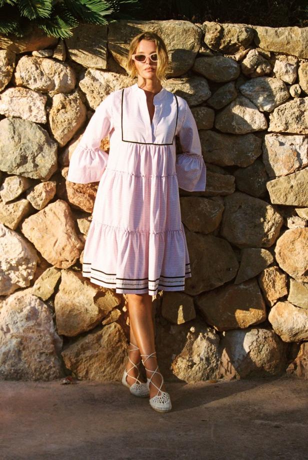 Eine Frau in einem rosa-weiß gestreiften Kleid und weißen Sandalen posiert vor einer Steinmauer.