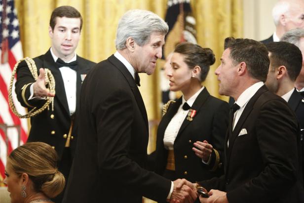 John Kerry schüttelt einem Mann die Hand, während Marineangehörige im Hintergrund stehen.