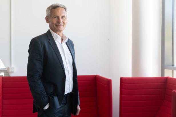 Ein Mann in Anzug und Hemd steht vor roten Sitzelementen.