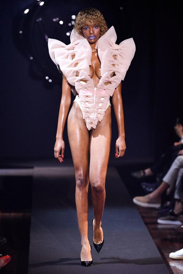 Eine Frau mit lila Lippenstift trägt ein extravagantes, schleifenverziertes Outfit auf dem Laufsteg.