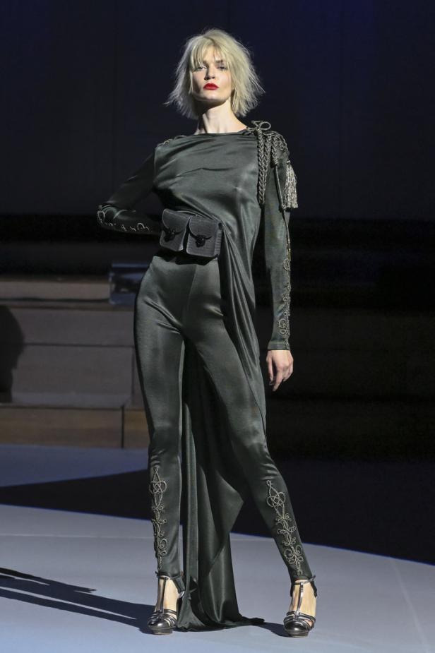 Ein Model präsentiert ein olivgrünes, eng anliegendes Kleid mit Ziertasche und Stickereien auf dem Laufsteg.