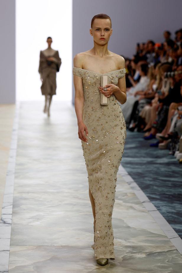 Ein Model präsentiert ein schulterfreies, beiges Kleid mit Pailletten auf einem Laufsteg.