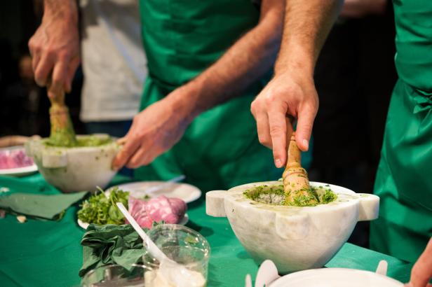 Zwei Personen in grünen Schürzen bereiten Pesto mit Mörser und Stößel zu.
