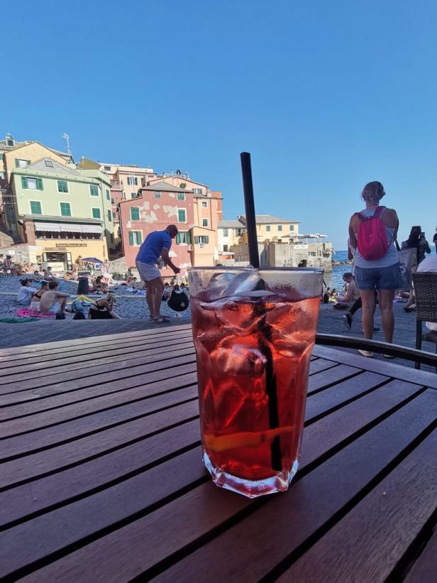 Ein roter Cocktail mit Eiswürfeln auf einem Tisch, im Hintergrund ein Strand mit Menschen und bunten Häusern.