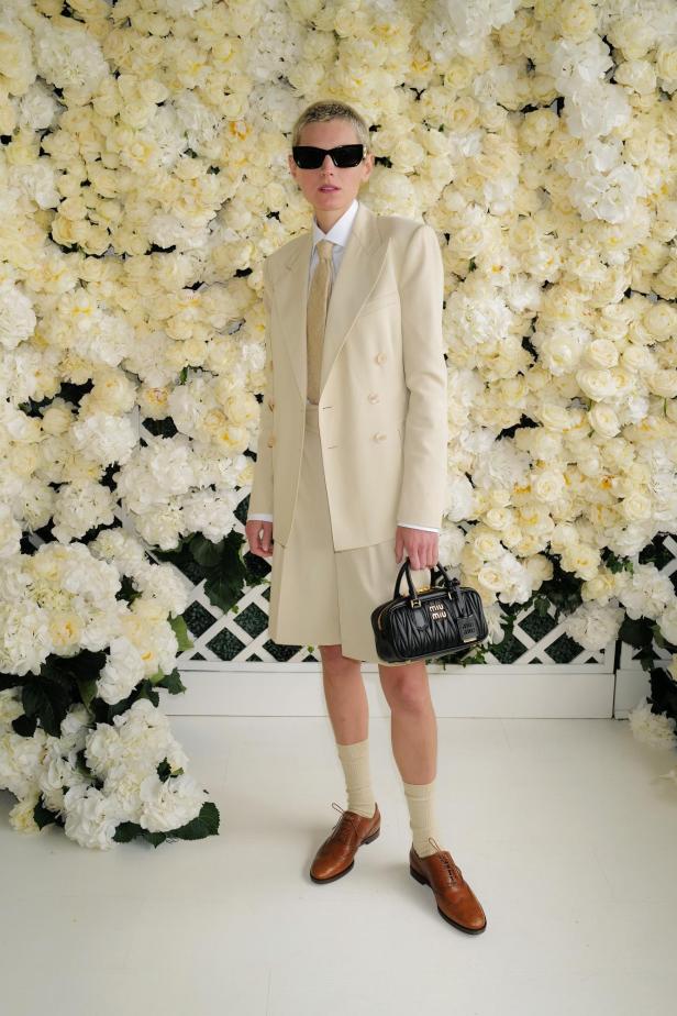 Eine Person mit Sonnenbrille trägt einen beigen Anzug und eine schwarze Miu Miu Handtasche vor einer Blumenwand.