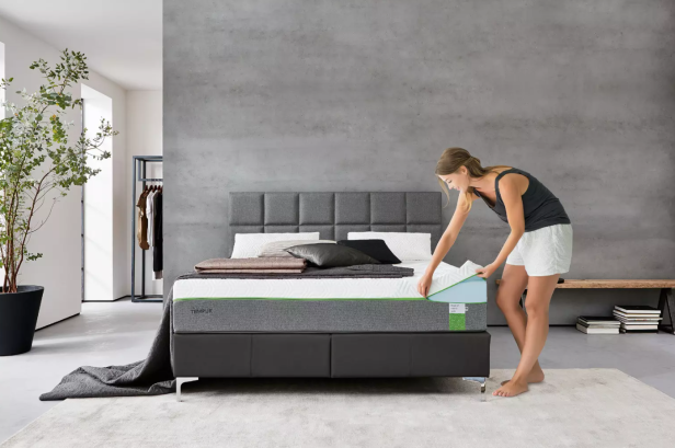 Eine Frau bezieht ein modernes Bett mit grauem Kopfteil in einem minimalistisch eingerichteten Schlafzimmer.