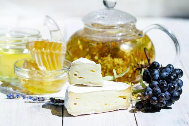 Brie type of cheese. Camembert cheese. Close up. Fresh Brie cheese and a slice on a wooden white table with honey, black grapes, herbal tea and lavender. Italian, French cheese.