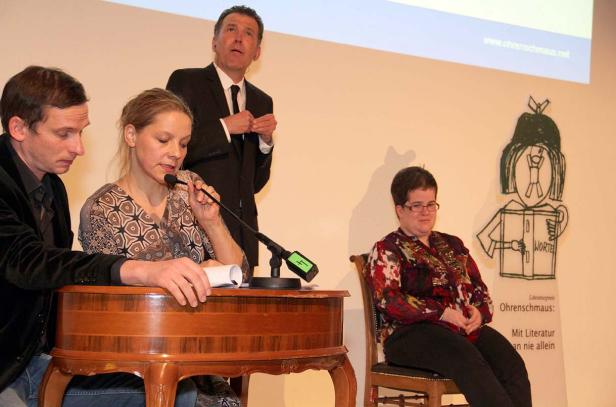 Eine Podiumsdiskussion mit drei Personen und einer Zuhörerin vor einem Logo von Ohrenschmaus.