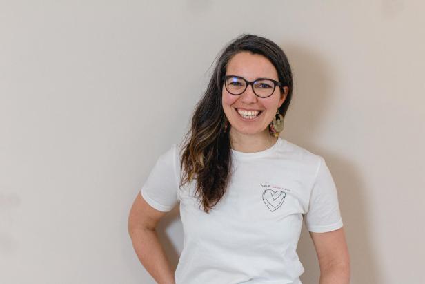 Eine lächelnde Frau mit Brille und einem T-Shirt mit der Aufschrift „Self Love Club“.