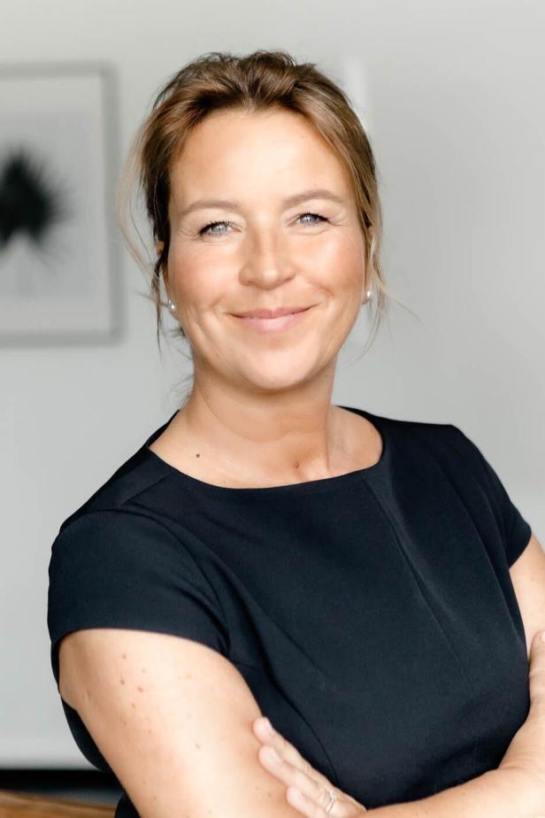 Eine lächelnde Frau mit blonden Haaren und einem schwarzen Kleid vor einem hellen Hintergrund.