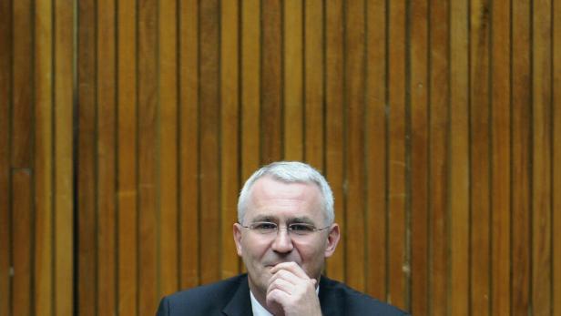 Ein Mann mit Brille und grauem Haar vor einer Holzwand hält seine Hand ans Kinn.