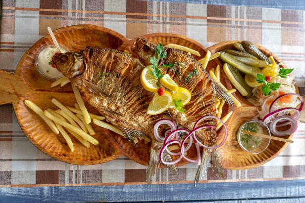 Zwei gebratene Fische werden mit Pommes, Gurken und Zwiebeln auf einem Holzbrett serviert.