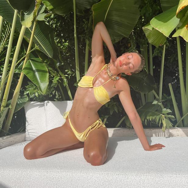 Eine Frau in einem gelben Bikini dehnt sich auf einer weißen Liege vor tropischen Pflanzen.