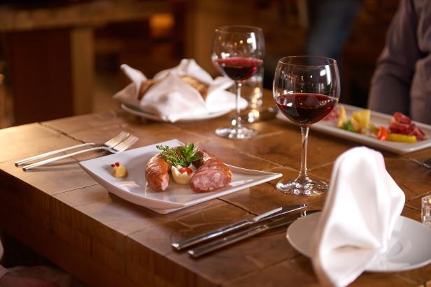 Auf einem Holztisch steht eine Wurst mit Kräutern und Senf auf einem Teller, dazu Rotwein.