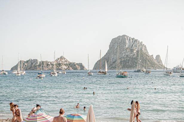 Menschen entspannen an einem Strand auf Ibiza mit Blick auf das Meer und Segelboote.