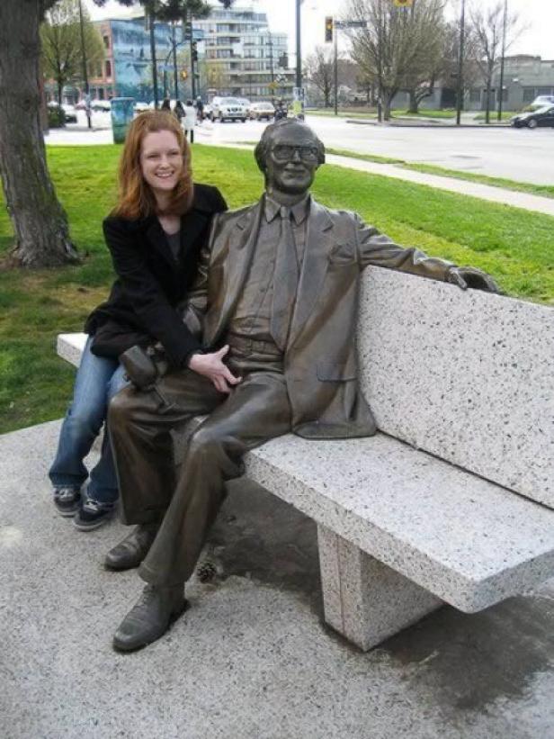 Eine Frau sitzt neben einer Bronzestatue auf einer Bank in einem Park.
