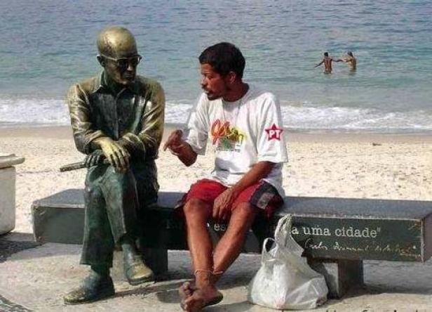 Ein Mann sitzt neben einer Statue von Carlos Drummond de Andrade an einem Strand.