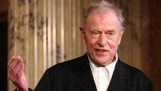 Ein Mann in einem schwarzen Anzug gestikuliert vor einem gemusterten Hintergrund.