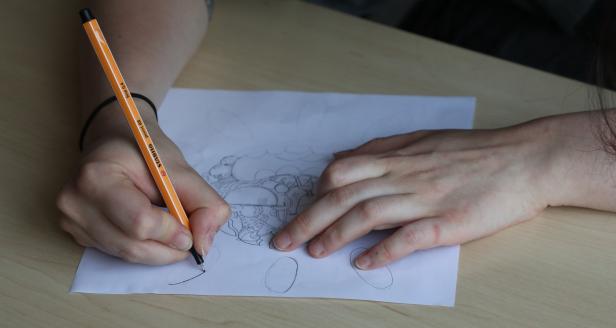 Eine Person zeichnet mit einem orangefarbenen Stift auf einem Blatt Papier.