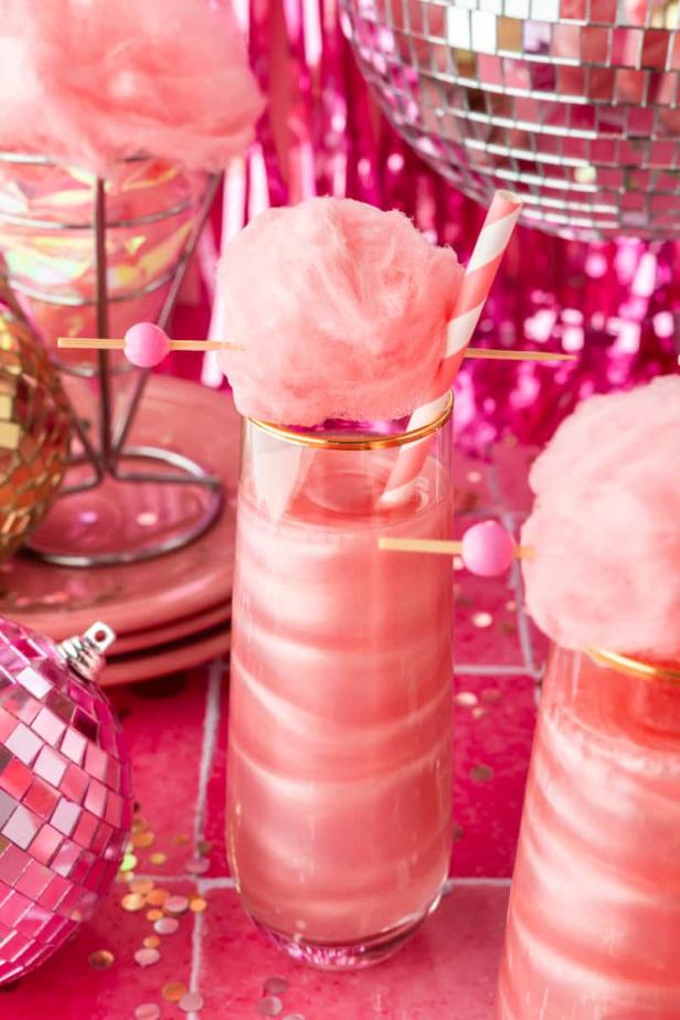Ein rosa Cocktail mit Zuckerwatte und einer gestreiften Papierstrohhalm vor einer glitzernden Dekoration.