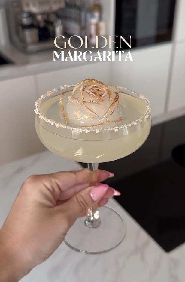 Ein Cocktail mit einem Eiswürfel in Form einer Rose und goldenen Akzenten wird von einer Hand gehalten.