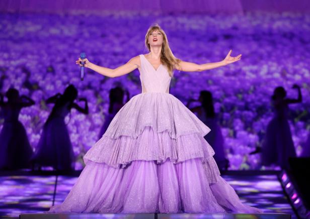 Taylor Swift tritt in einem lavendelfarbenen Kleid auf einer Bühne vor einem Blumenhintergrund auf.