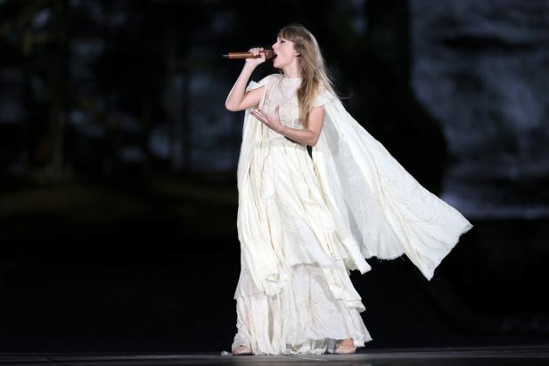 Taylor Swift singt in einem langen, weißen Kleid mit Cape auf einer Bühne.