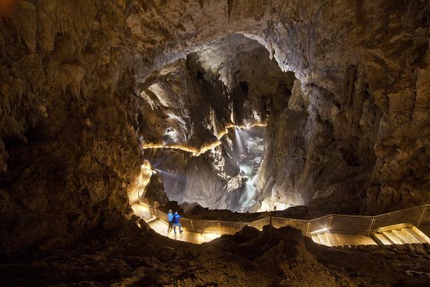 Ein Mann steht auf einem Steg in einer beleuchteten Höhle mit einem Wasserfall im Hintergrund.