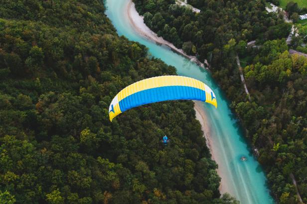 Ein Gleitschirmflieger fliegt über einen türkisfarbenen Fluss, umgeben von dichtem Wald.