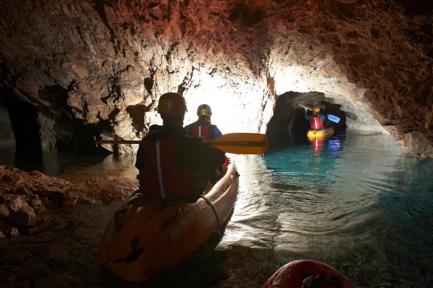 Personen paddeln in Kajaks durch eine beleuchtete Höhle mit türkisfarbenem Wasser.