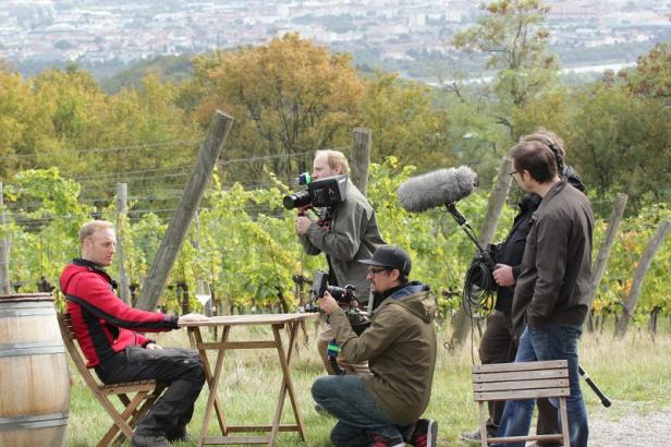 Ein Mann sitzt an einem Tisch in einem Weinberg, während ein Filmteam ihn filmt.
