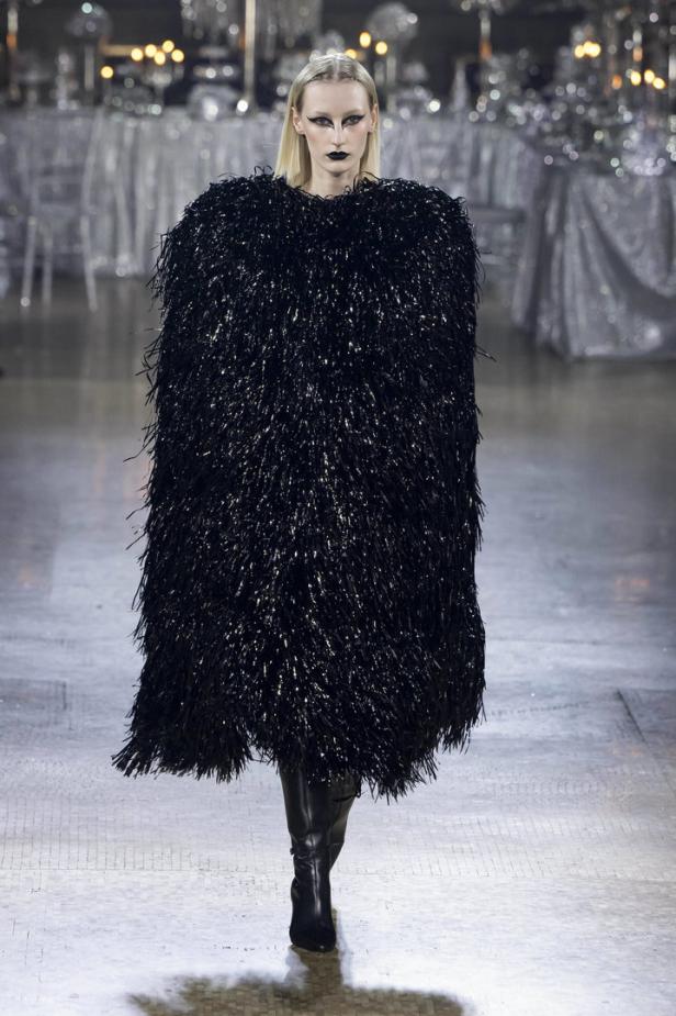 Ein Model mit dunklem Make-up trägt ein schimmerndes, schwarzes Kleid auf dem Laufsteg.