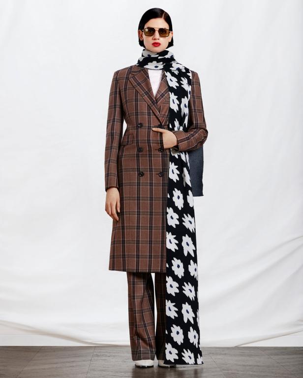 Eine Frau in einem braunen Hosenanzug mit Karomuster und einem schwarz-weißen Schal mit Blumenmuster.