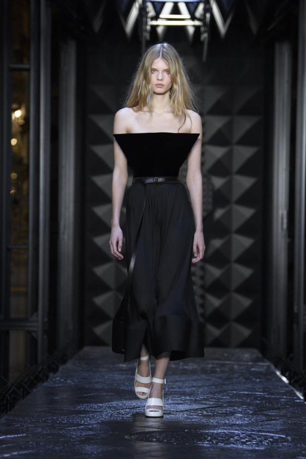 Eine Frau mit blonden Haaren läuft in einem schwarzen Kleid und weißen Sandalen über einen Laufsteg.