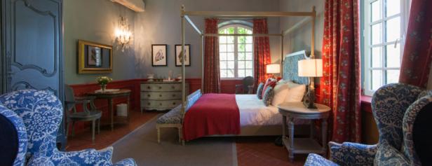 Ein Schlafzimmer mit Himmelbett, roten Vorhängen und blau-weiß gemusterten Sesseln.