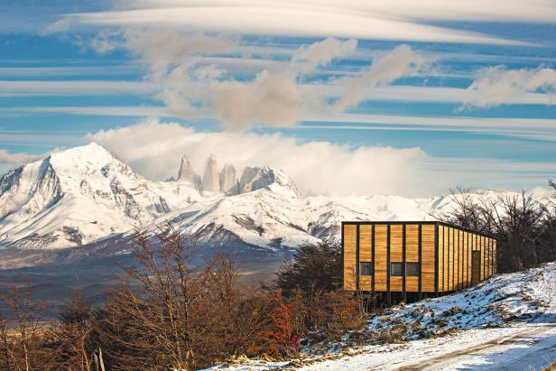Ein modernes Holzhaus vor einer schneebedeckten Berglandschaft mit den Torres del Paine im Hintergrund.