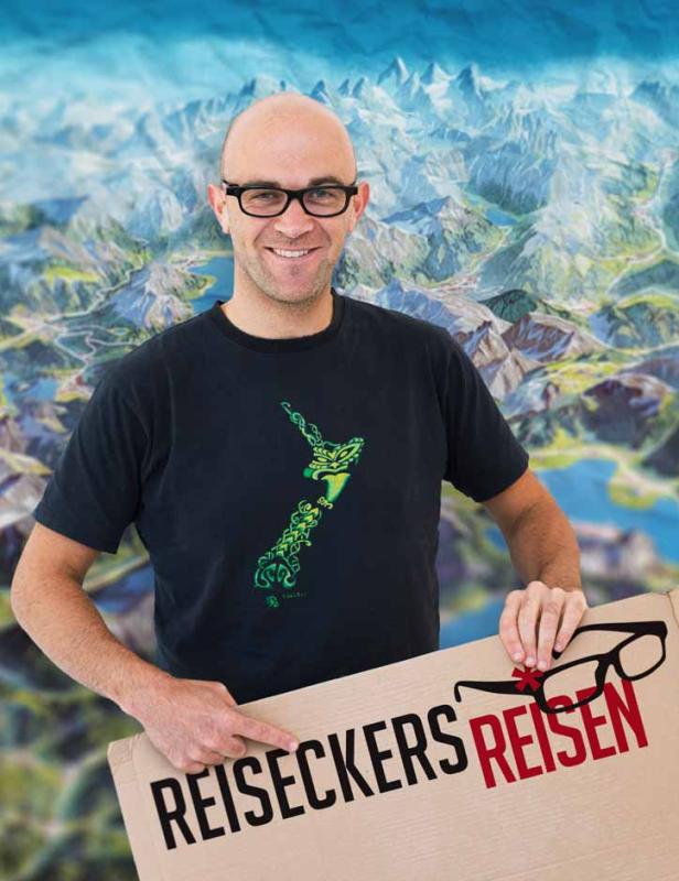 Ein Mann mit Brille präsentiert ein Schild mit der Aufschrift „Reiseckers Reisen“ vor einer Bergkulisse.