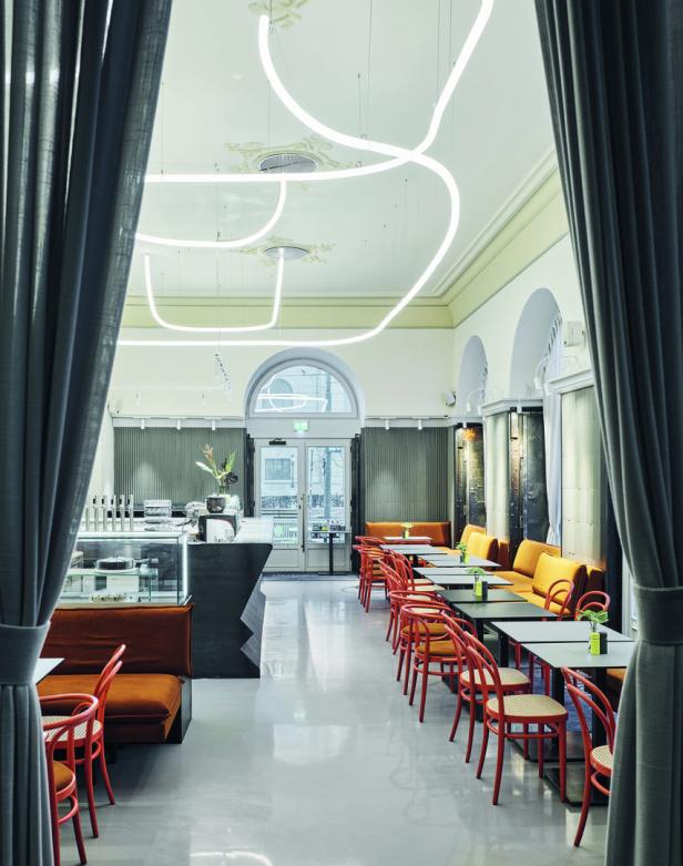 Innenansicht eines Cafés mit orangefarbenen Stühlen und Sofas unter geschwungenen Neonleuchten.