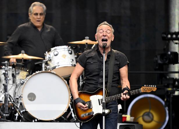Bruce Springsteen singt und spielt Gitarre auf der Bühne, während ein Schlagzeuger im Hintergrund zu sehen ist.