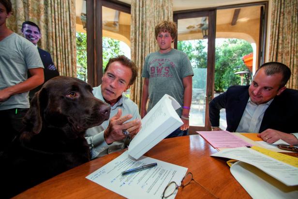 Arnold Schwarzenegger sitzt mit einem braunen Labrador und anderen Personen an einem Tisch voller Dokumente.
