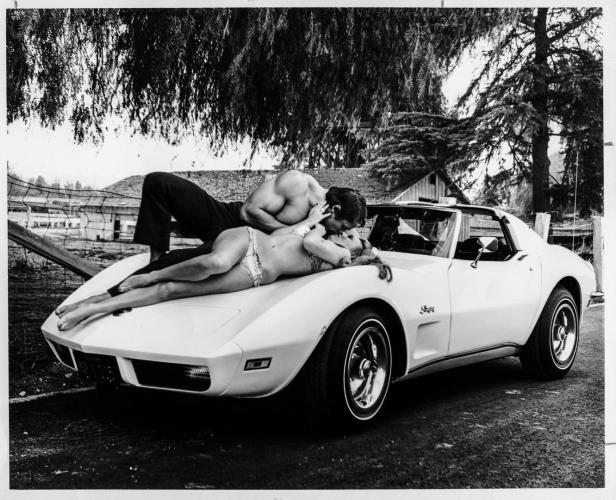 Ein Paar liegt auf der Motorhaube eines weißen Sportwagens und küsst sich.