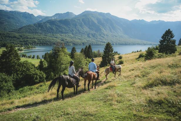 Drei Personen reiten auf Pferden vor einer Bergkulisse und einem See.