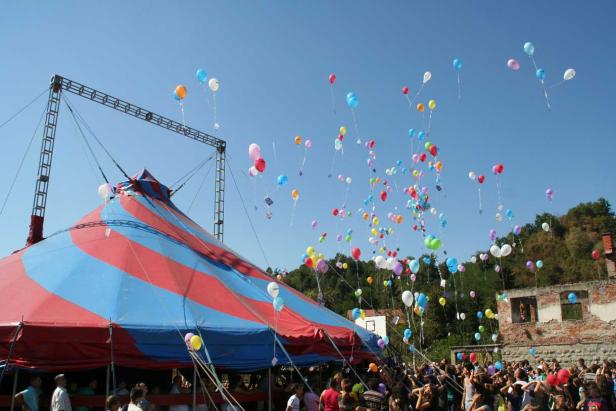 Vor einem Zirkuszelt steigen viele bunte Ballons in den Himmel auf.