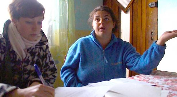 Zwei Frauen sitzen an einem Tisch; eine schreibt, die andere gestikuliert erklärend.