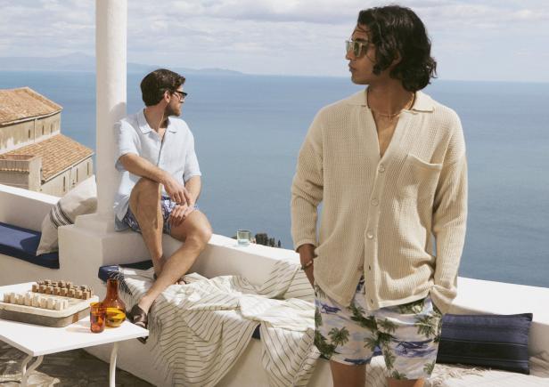 Zwei Männer in Sommerkleidung entspannen auf einer Terrasse mit Blick auf das Meer.