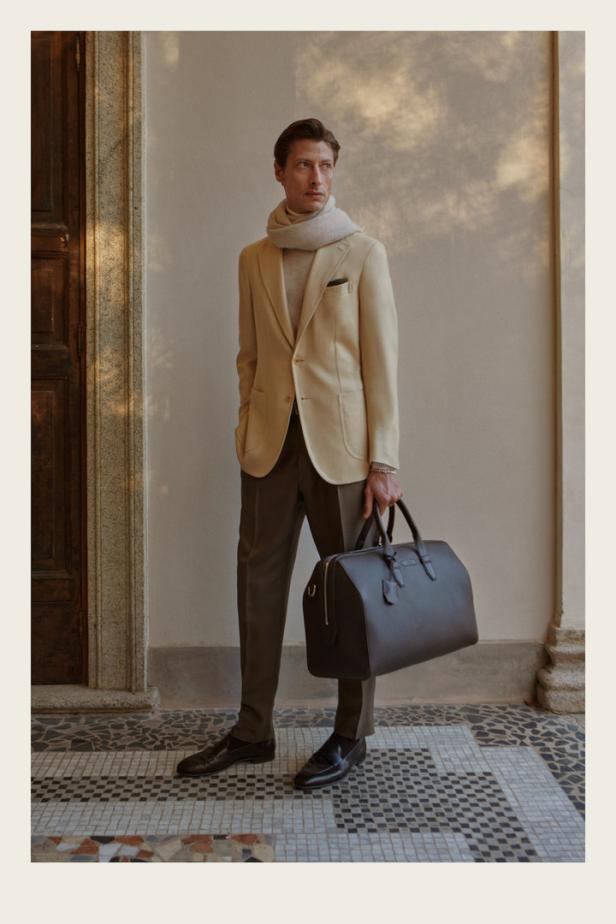 Ein Mann in einem beigen Sakko und brauner Hose hält eine Reisetasche.