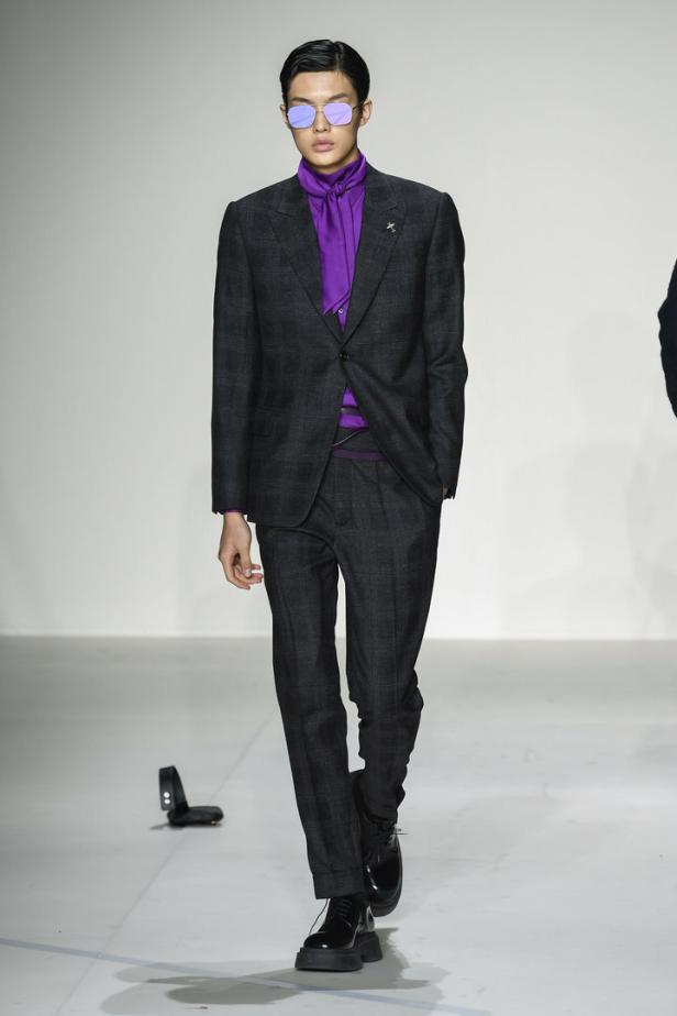 Ein Mann mit Sonnenbrille trägt einen grauen Anzug und ein violettes Hemd auf dem Laufsteg.